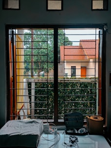A window view displaying a house with a red-tiled roof and green trees in the background. In the foreground, there is a table with electronic cords, a power strip, and a rolled-up yoga mat. The window has metal bars and light curtains hanging to the side.