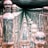 An arrangement of various glass bottles and containers is displayed, featuring a repeated reflection effect that creates a seemingly infinite corridor of objects. The glass is transparent and highly reflective, capturing light and surrounding colors, resulting in a distorted yet mesmerizing visual. The objects vary in shape, including spherical, cylindrical, and elongated designs.