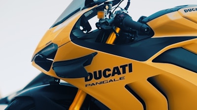 A close-up shot of a bright yellow Ducati Panigale motorcycle, showcasing its sleek design and sport features. The brand and model name are prominently displayed on the side of the bike. The handlebar and part of the windscreen are visible, highlighting its aerodynamic structure.