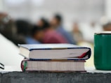Two books with colorful bindings are stacked on a rough-textured surface, alongside a green cup. In the background, several blurred figures, suggesting a classroom or seminar setting, are visible, adding depth to the scene.