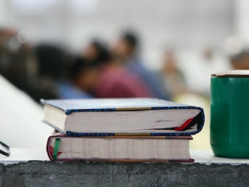 Two books with colorful bindings are stacked on a rough-textured surface, alongside a green cup. In the background, several blurred figures, suggesting a classroom or seminar setting, are visible, adding depth to the scene.
