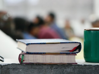 Two books with colorful bindings are stacked on a rough-textured surface, alongside a green cup. In the background, several blurred figures, suggesting a classroom or seminar setting, are visible, adding depth to the scene.