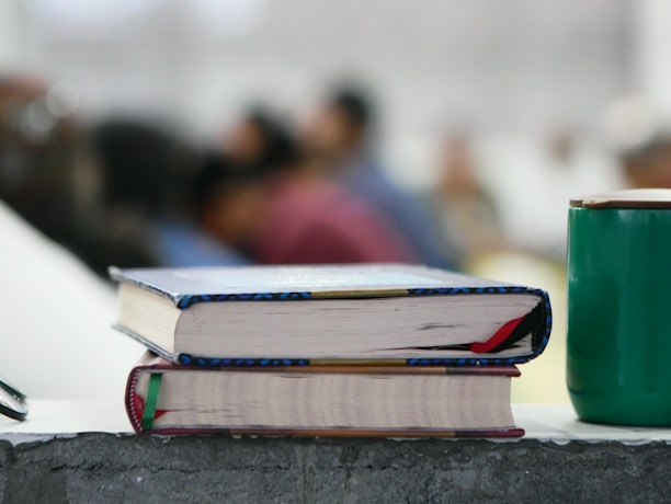Two books with colorful bindings are stacked on a rough-textured surface, alongside a green cup. In the background, several blurred figures, suggesting a classroom or seminar setting, are visible, adding depth to the scene.