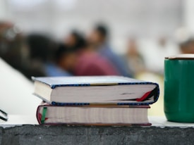 Two books with colorful bindings are stacked on a rough-textured surface, alongside a green cup. In the background, several blurred figures, suggesting a classroom or seminar setting, are visible, adding depth to the scene.