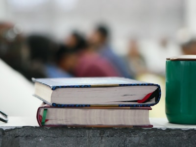 Two books with colorful bindings are stacked on a rough-textured surface, alongside a green cup. In the background, several blurred figures, suggesting a classroom or seminar setting, are visible, adding depth to the scene.