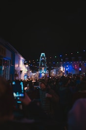 A bustling street scene during a nighttime event, with a crowd of people gathered under festive string lights of various colors. Buildings on either side reflect the colorful illumination, creating a lively and vibrant atmosphere. Many attendees are taking photos, capturing the festive moment.