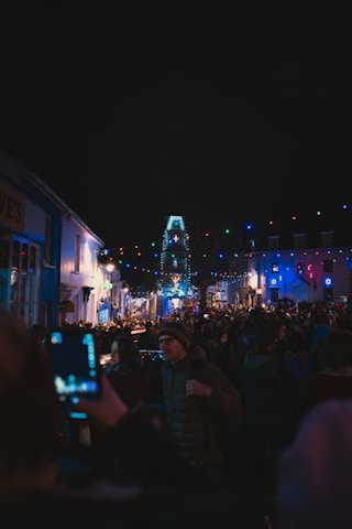 A bustling street scene during a nighttime event, with a crowd of people gathered under festive string lights of various colors. Buildings on either side reflect the colorful illumination, creating a lively and vibrant atmosphere. Many attendees are taking photos, capturing the festive moment.