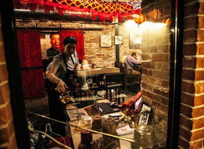 A cozy, dimly lit restaurant with exposed brick walls and red Chinese-style lanterns hanging from the ceiling. A person is pouring tea at a table, while a few patrons are seated, one of whom is working on a laptop. Chinese artwork and a large portrait of a person hang on the walls, adding to the cultural ambiance.