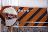 An orange construction sign with black diagonal stripes is next to a circular convex mirror. The sign contains white text in a non-Latin script, and the scene appears to be part of an industrial or construction area, with protective barriers visible in the background.