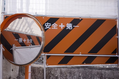 An orange construction sign with black diagonal stripes is next to a circular convex mirror. The sign contains white text in a non-Latin script, and the scene appears to be part of an industrial or construction area, with protective barriers visible in the background.