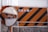 An orange construction sign with black diagonal stripes is next to a circular convex mirror. The sign contains white text in a non-Latin script, and the scene appears to be part of an industrial or construction area, with protective barriers visible in the background.
