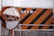An orange construction sign with black diagonal stripes is next to a circular convex mirror. The sign contains white text in a non-Latin script, and the scene appears to be part of an industrial or construction area, with protective barriers visible in the background.