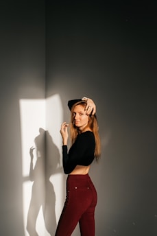 A woman stands confidently in a well-lit space, with sunlight casting her shadow on the wall. She is wearing a black long-sleeve top and burgundy pants, posing elegantly with one hand raised near her head.