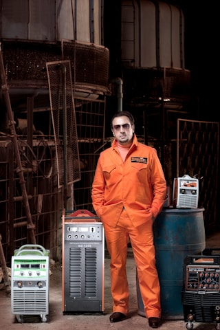 A man wearing an orange jumpsuit stands with his hands in his pockets, surrounded by industrial equipment. The setting is an industrial area with metal structures and machinery in the background. Various pieces of equipment, including welding machines or similar devices, are positioned around him, some on a blue barrel.