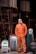 A man wearing an orange jumpsuit stands with his hands in his pockets, surrounded by industrial equipment. The setting is an industrial area with metal structures and machinery in the background. Various pieces of equipment, including welding machines or similar devices, are positioned around him, some on a blue barrel.