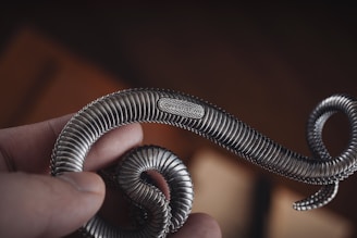 A hand holding a metallic, intricately designed snake sculpture with detailed textures and an engraved label reading 'COPPERTIST.WU'. The sculpture's surface features ribbed segments and loops in a twisting form.