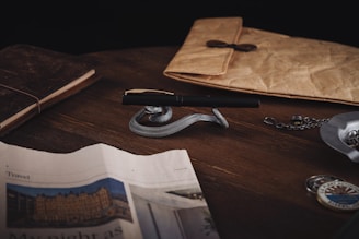 A wooden table holds a luxurious pen resting on a silver snake-shaped holder. Nearby, a leather-bound notebook and a rustic envelope with a button closure are placed. A newspaper partially covers the table, and a pocket watch on a chain lies to the right. The scene conveys a vintage, sophisticated ambiance.