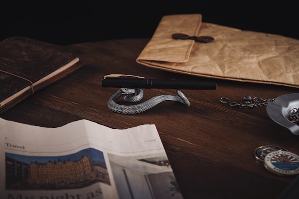 A wooden table holds a luxurious pen resting on a silver snake-shaped holder. Nearby, a leather-bound notebook and a rustic envelope with a button closure are placed. A newspaper partially covers the table, and a pocket watch on a chain lies to the right. The scene conveys a vintage, sophisticated ambiance.