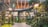 A modern indoor garden features lush green foliage, tall trees, and decorative street lamps. The area is illuminated with warm lighting and surrounded by a multi-level shopping mall structure with colorful signage in the background.