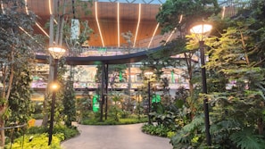 A modern indoor garden features lush green foliage, tall trees, and decorative street lamps. The area is illuminated with warm lighting and surrounded by a multi-level shopping mall structure with colorful signage in the background.