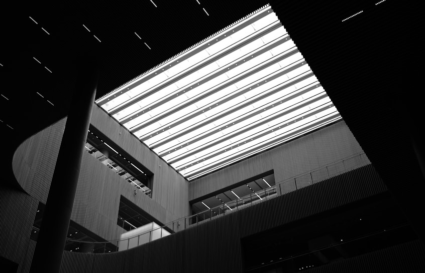 A high contrast black and white interior view showcasing a modern architectural design with a large rectangular skylight. The space features tall vertical columns and an array of horizontal lines created by slatted paneling. Geometric shapes and shadows play on the surfaces, adding depth and dimension.