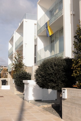 Modern apartment buildings with glass balconies and a yellow and blue flag hanging from one of the apartments. The area is landscaped with neatly trimmed bushes and trees, and there are paved walkways. The architecture is clean with white facades.