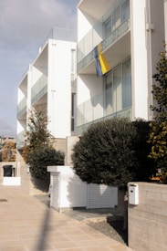Modern apartment buildings with glass balconies and a yellow and blue flag hanging from one of the apartments. The area is landscaped with neatly trimmed bushes and trees, and there are paved walkways. The architecture is clean with white facades.