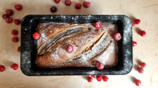 A freshly baked loaf of banana bread rests in a black rectangular pan. The top of the bread is sprinkled with powdered sugar and decorated with whole cranberries. Surrounding the baking pan on the countertop are more cranberries, adding to the rustic and homemade feel.
