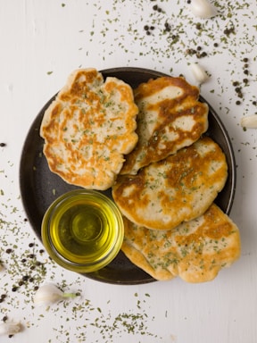 A dark plate holds four pieces of flatbread with a golden-brown crust, sprinkled with herbs. Beside the bread, a small clear bowl contains olive oil. Scattered around the plate are herbs, garlic cloves, and peppercorns on a light wooden surface.