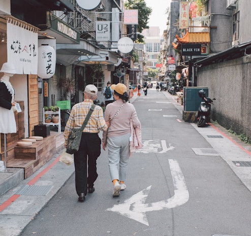 An elderly couple walks arm-in-arm down a narrow, urban street lined with small shops and cafes. The street is decorated with various signs in different languages, and there are occasional pedestrians and a parked scooter. The atmosphere appears calm and leisurely.