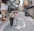 An elderly couple walks arm-in-arm down a narrow, urban street lined with small shops and cafes. The street is decorated with various signs in different languages, and there are occasional pedestrians and a parked scooter. The atmosphere appears calm and leisurely.