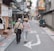 An elderly couple walks arm-in-arm down a narrow, urban street lined with small shops and cafes. The street is decorated with various signs in different languages, and there are occasional pedestrians and a parked scooter. The atmosphere appears calm and leisurely.