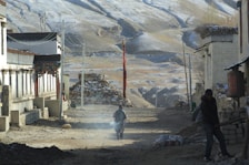 A narrow, dusty street lined with traditional stone and adobe buildings, with a person walking in the middle. The background features a series of barren, snow-dusted hills, and the scene is framed by a bright red vertical flag and a small stone structure.