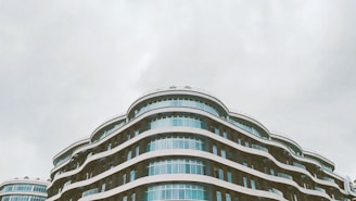 A modern architectural building featuring multiple curved layers and large glass windows stands against a cloudy sky. The design includes flowing lines and a sleek, contemporary aesthetic.