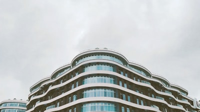A modern architectural building featuring multiple curved layers and large glass windows stands against a cloudy sky. The design includes flowing lines and a sleek, contemporary aesthetic.