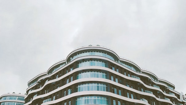A modern architectural building featuring multiple curved layers and large glass windows stands against a cloudy sky. The design includes flowing lines and a sleek, contemporary aesthetic.