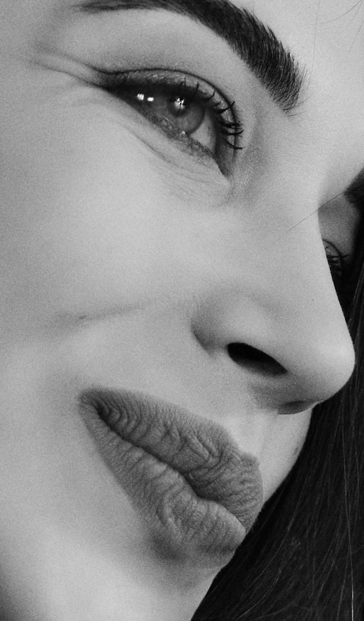 A close-up black and white portrait focuses on the side of a person's face, highlighting the eye, eyebrow, and mouth. The image captures fine details such as eyelashes, skin texture, and subtle lighting.