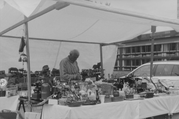 An elderly person is standing under a canopy, attending to a variety of vintage items displayed on tables. The scene is set in an outdoor market, with a car parked nearby. The collection includes miniature models, ornate boxes, and various knick-knacks.
