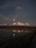 A tranquil landscape features a calm water body reflecting a bright moon and fluffy clouds. The horizon displays an open field with a backdrop of mountain silhouettes under a darkening sky. The scene is bordered by lush greenery and a clear reflection of the sky in the water, adding to the serene ambiance.