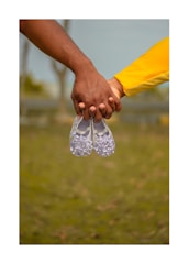 Two hands are holding a pair of small, white shoes, suggesting a focus on themes such as family or expectancy. The hands are of different skin tones, and one sleeve is visible in bright yellow. The background is blurred, showing an outdoor setting with green grass and trees.