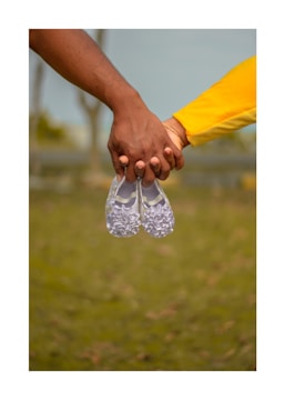 Two hands are holding a pair of small, white shoes, suggesting a focus on themes such as family or expectancy. The hands are of different skin tones, and one sleeve is visible in bright yellow. The background is blurred, showing an outdoor setting with green grass and trees.