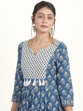 A woman wearing a blue patterned traditional dress with white embroidery and designs. Her hair is tied back, and she is accessorized with long, ornate earrings. The dress features a white and blue patterned yoke and tassel details.