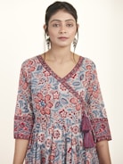A woman is wearing a floral patterned dress with intricate designs in shades of blue, red, and pink. The dress features a V-neckline and 3/4 sleeves with detailed borders. She accessorizes with large dangling earrings and has her hair styled neatly. The background is plain and light-colored, enhancing the focus on the attire.