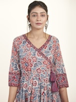 A woman is wearing a floral patterned dress with intricate designs in shades of blue, red, and pink. The dress features a V-neckline and 3/4 sleeves with detailed borders. She accessorizes with large dangling earrings and has her hair styled neatly. The background is plain and light-colored, enhancing the focus on the attire.