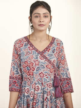 A woman is wearing a floral patterned dress with intricate designs in shades of blue, red, and pink. The dress features a V-neckline and 3/4 sleeves with detailed borders. She accessorizes with large dangling earrings and has her hair styled neatly. The background is plain and light-colored, enhancing the focus on the attire.