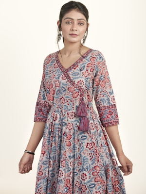 A woman is wearing a floral-patterned dress with red, blue, and white designs. The dress features three-quarter length sleeves and a V-neckline with tassels. She has earrings, makeup, and her hair is styled back.