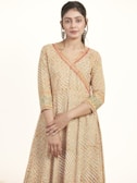 A person is wearing a traditional, patterned dress that features intricate embroidery with a mix of colors, including orange, green, and white. Their hands are gently clasped in front, and they are wearing large, ornate hoop earrings. The background is plain and light, highlighting the detailed patterns of the attire.