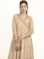 A person is wearing a traditional, patterned dress that features intricate embroidery with a mix of colors, including orange, green, and white. Their hands are gently clasped in front, and they are wearing large, ornate hoop earrings. The background is plain and light, highlighting the detailed patterns of the attire.