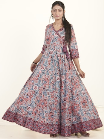 A woman is wearing a long, flowing dress with an intricate floral pattern. The dress features a mix of red, blue, and white colors and has three-quarter sleeves. She has long, dark hair and is accessorized with earrings and bracelets. The background is a soft, neutral tone, allowing the dress to stand out.