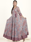 A woman wearing a long, flowing dress with a detailed floral pattern in shades of red, blue, and white. The dress has three-quarter sleeves and a V-neckline, complemented by a tassel detail at the waist. She has long hair styled straight and is wearing earrings. The background is plain and light-colored.